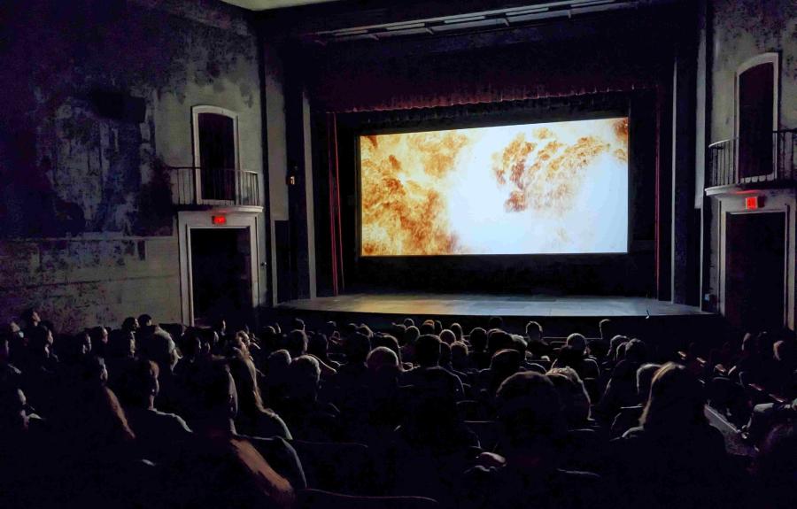An audience sits in the dark looking at a movie screen filled with a fiery image from Christopher Nolan's Oppenheimer (2023).