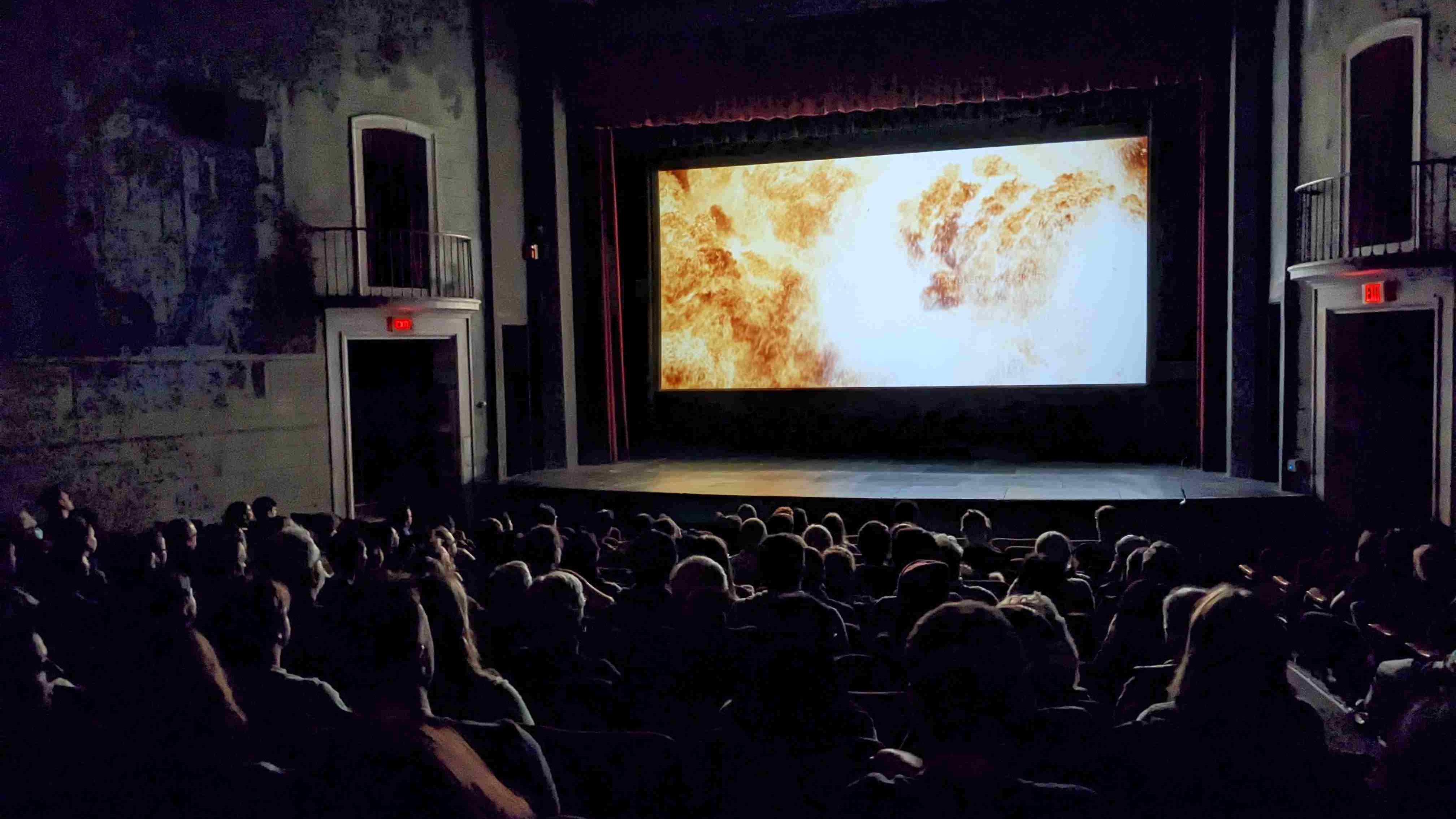 An audience sits in the dark looking at a movie screen filled with a fiery image from Christopher Nolan's Oppenheimer (2023).
