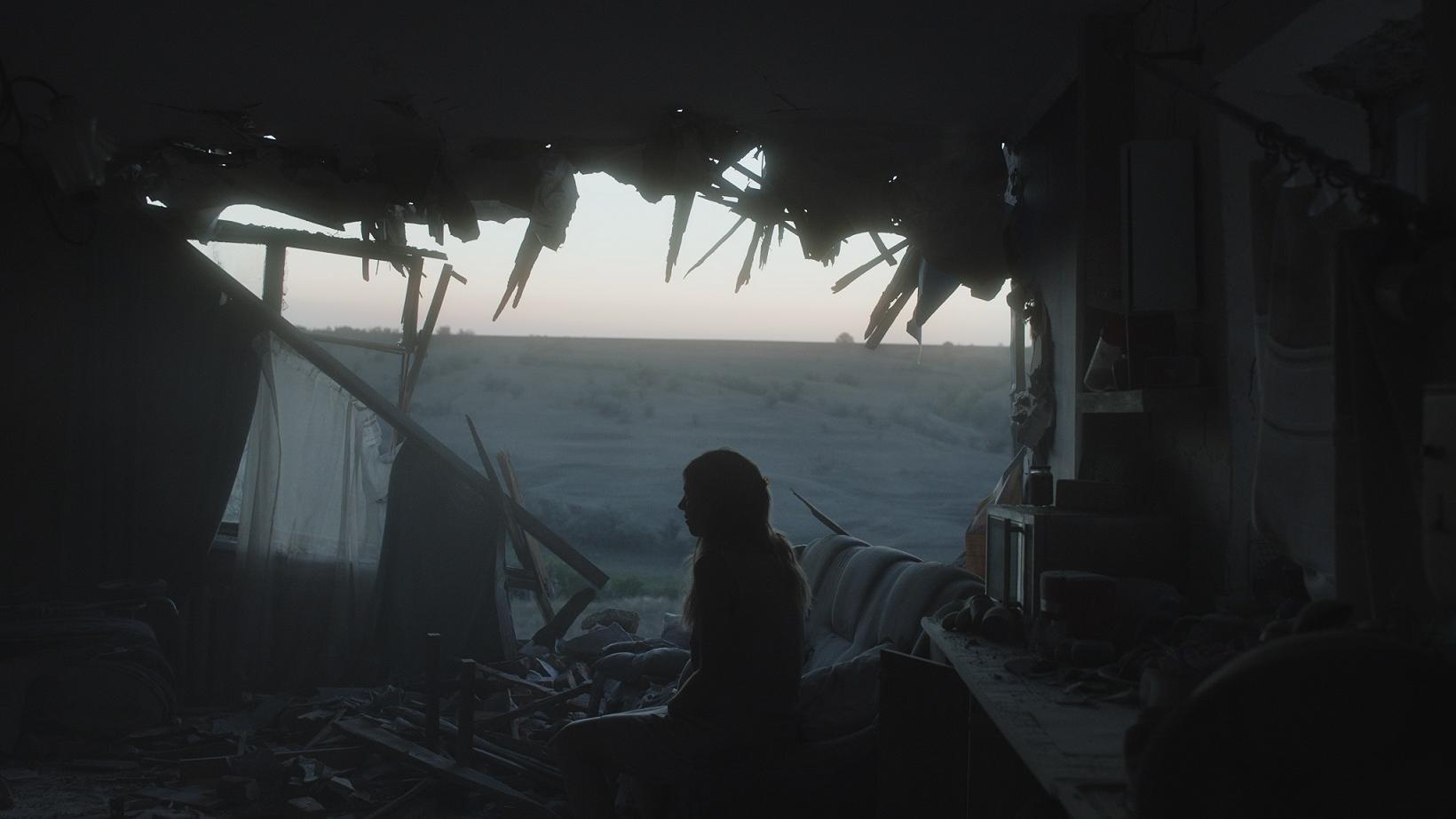 A woman sitting next to the destroyed wall of a house.