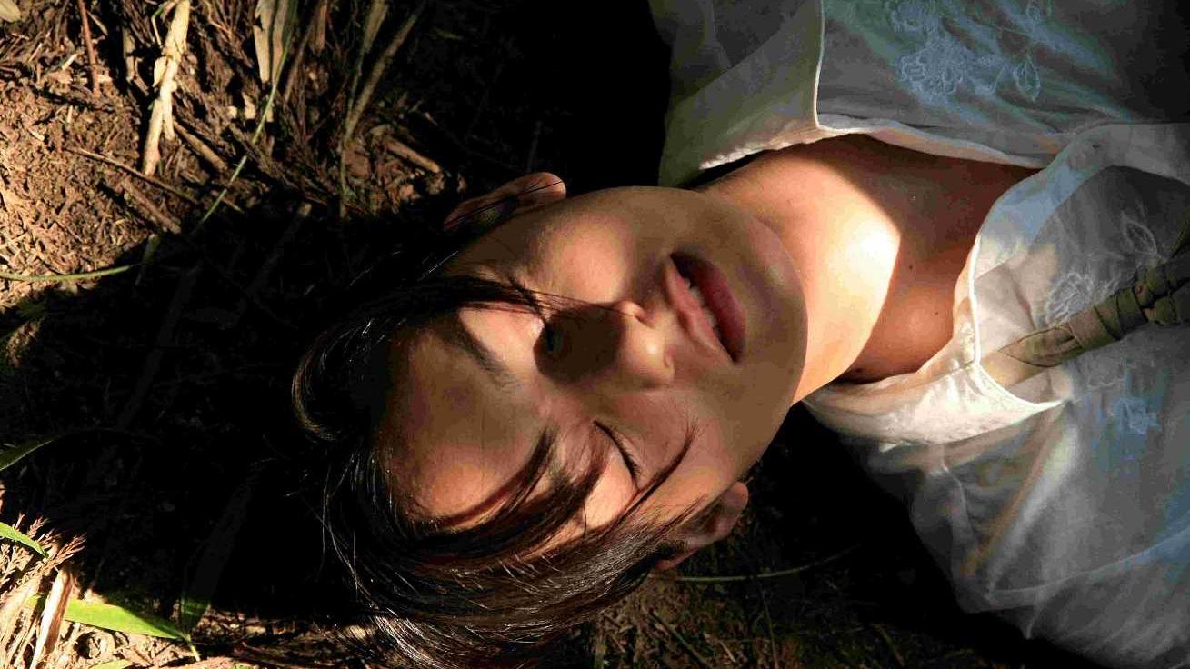 A woman in dappled sunlight laying on the ground with her eyes closed.