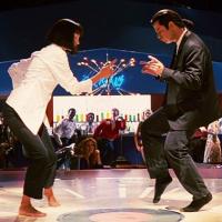 A woman in a white shirt and a man in a black suit dance the twist at the center of a night club/restaurant dance floor.