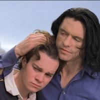 A young boy rests his head tenderly on the shoulder of a man with long dark hair who embraces him in return.