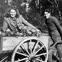 A black-and-white photograph of a yougng woman sitting in a wheel barrow being pushed by a young man.