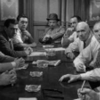 A black and white photograph of eleven individuals sitting around a long table. The setting appears to be a formal meeting or discussion, with most individuals wearing suits and ties. Several documents or papers are visible on the table in front of them.