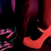 Close-up of a person’s lower legs and feet. One foot is wearing a high-heeled shoe, while the other foot is bare and stepping on a piece of clothing or fabric on the floor. The lighting has a reddish-purple hue, adding a dramatic effect to the scene.