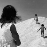 Black and white photograph of three individuals, likely children, on a mound of earth or sand. The foreground shows the back of one individual with long hair, looking towards the other two who are further up the mound. The sky is clear and bright, suggest