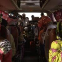 Interior of a crowded bus with passengers seated facing forward. The bus appears to be in motion, and the passengers are dressed in various colorful garments. Luggage is stored above the seats, and the windows are covered with curtains. The atmosphere see