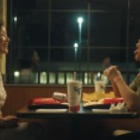 Two individuals seated opposite each other at a table inside a fast-food restaurant at night. On the table, there are various fast-food items including a drink with a straw, food wrappers, and partially eaten meals. The bright signage of McDonald’s is vis