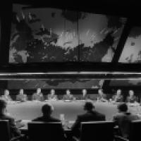 A black and white photo of a group of men seated around a large, oval-shaped table in a room with dim lighting. Above the table hangs a circular light fixture that illuminates the area directly below. Behind the men is an expansive wall map displaying var