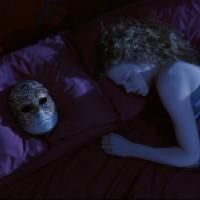 A person with light-colored hair is lying on a bed with purple bedding, appearing to be asleep. Next to the person is an ornate mask with gold detailing and white feathers on a dark background. The scene has a nocturnal ambiance, suggesting it could be ni