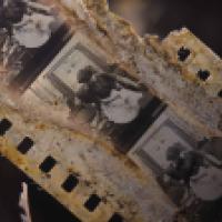 An old, deteriorated film strip with a series of black and white images. The film strip has visible damage and rust, indicating age and decay. The images on the film appear to be from an early 20th-century performance or play, featuring individuals in per