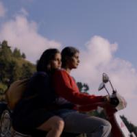 Two individuals on a scooter are stationary with a backdrop of a hill covered in greenery under a clear sky. The person in the front appears to be controlling the scooter while the one at the back is sitting close, possibly holding onto the driver or the 