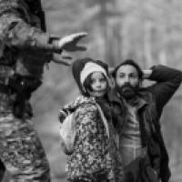 A black and while photo of a distressed man with his hands behind his head, kneeling next to a child, with a solider in army fatigues and a large gun in the foreground.