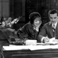 A black and white photograph featuring two individuals sitting at a desk. One individual appears to be reclining with legs crossed on the desk, while the other is seated upright, seemingly writing or signing a document. On the desk, there are various item