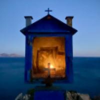 A small shrine or religious icon display with a Christian cross on top. It is set against a blurred background featuring what appears to be a body of water and possibly distant land or mountains. The shrine is illuminated from within, casting a warm glow,