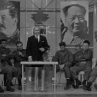 A black and white photograph featuring multiple individuals seated in a row. Standing behind the seated individuals is another figure, also with their face obscured, positioned next to a table with what appears to be a microphone on it. The background inc