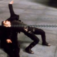 A scene from a film with two characters in the midst of an action sequence. One character is dressed in black and is dodging backward with an exaggerated arch in their back, while the other character, also dressed in black, appears to be shooting at the f