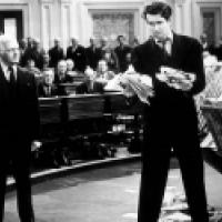 A black and white photograph showing a scene with multiple individuals in what appears to be a legislative assembly or courtroom. One of these individuals is standing at a podium, holding papers, and appears to be addressing the assembly, while the other 