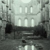 The ruins of a large gothic structure with tall arches and no roof. In the foreground, there is a small body of water reflecting part of the structure. A person is seated near this water, and there are several other individuals grouped together slightly f