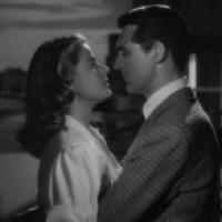 A black and white photograph featuring two individuals facing each other closely as if they are about to kiss. The setting appears to be indoors with a softly focused background that suggests a domestic or intimate environment.