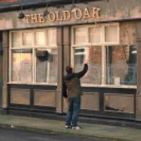 An individual standing in front of a building with a sign that reads “THE OLD OAK.” The person appears to be reaching up towards one of the crooked sign letters with a stick or tool to fix it. The building has a traditional facade, suggesting it might be 