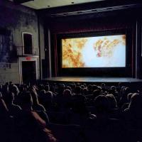 An audience sits in the dark looking at a movie screen filled with a fiery image from Christopher Nolan's Oppenheimer (2023).