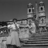 A black and white photograph of a scene with multiple individuals on and around a large set of stone steps leading up to a building with two bell towers. The foreground features an individual standing on the steps. Another prominent figure is leaning agai