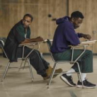 Two individuals seated on chairs at separate desks in a room with wooden paneling on the walls. Both individuals are wearing green pants and one is wearing a purple sweatshirt. The individual in the purple sweatshirt appears to be writing or drawing on a 