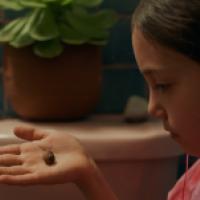 A seemingly young individual holding out their hand with a small snail on it. In the background, there is a potted plant sitting on what appears to be a bathroom sink or countertop.