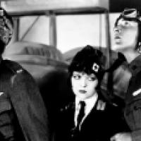 Three individuals dressed in military pilot uniforms standing in front of an aircraft. The uniforms have badges and insignia, suggesting a rank or affiliation with an air force. The setting appears historical due to the style of the uniforms and the desig