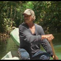 A man wearing a baseball hat rowing a boat on a lush river.