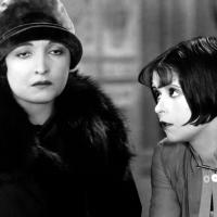 A close up shot of two women standing next to each other, one wearing a hat and one with a bobbed haircut.