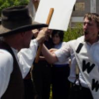 Men in 1917 period clothing gathered outside holding signs and shouting as part of a workers strike.