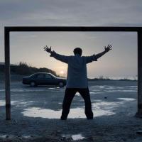 A man with arms outstretch standing in a square frame in an empty parkin lot next to the sea at dawn.