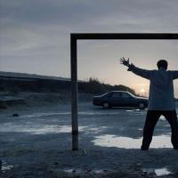 A man standing with his arms raised inside a square frame in an empty parking by the sea at dawn.