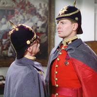 A shorter older solider in a gray uniform addresses a taller young soldier in a red uniform at close range.