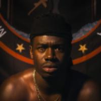 A person with a blurred face sits in front of a circular emblem with the text "MOVEMENT DELTA," featuring two gold stars and a graphic of a person in motion. The person is shirtless, wearing a headband and a gold chain, with dramatic lighting creating sha