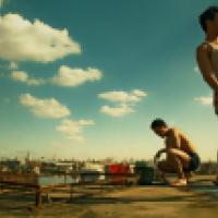 Two individuals are on a rooftop with a cityscape in the background. One stands in a white tank top and plaid shorts, while the other, shirtless, crouches nearby, surrounded by scattered objects like a rusted metal structure and a red bucket.