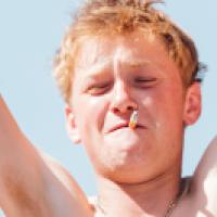 A person with a cigarette in mouth, arms raised, light brown or blonde hair, and a necklace stands against a clear blue sky background.