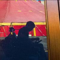 The reflection of a man in a motorized wheelchair with a red and yellow circus tent behind him.