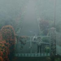 A gated entrance to a large garden or estate is visible in a foggy scene. The metal gate features decorative elements, and there is a person standing in front of it, possibly unlocking or opening it. Another person is seen behind the gate on a bicycle or 