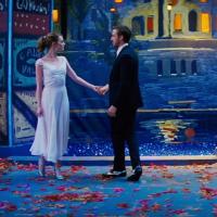 The image shows two people standing on a stage set that resembles a romantic, nighttime cityscape. The person on the left is wearing a white dress, and the person on the right is wearing a black suit. They are holding hands and facing each other. The background features a painted mural of a river with reflections of buildings and trees, illuminated by streetlights. The ground is covered with scattered red and orange leaves, and there are flowers on the right side of the image. The scene is visually striking