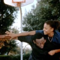 Two individuals are playing basketball outdoors. One individual is holding a basketball in an outstretched arm while the other is attempting to block or steal the ball. The basketball hoop and backboard are visible in the background, and the scene is set 