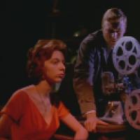 Two individuals are in a dimly lit room; one is seated and wearing a red dress, while the other is standing and operating a vintage film projector.