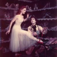 Two individuals in a theatrical setting; one stands in a light-colored dress while the other kneels, assisting with a red shoe, against a backdrop of shoe-filled shelves.