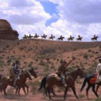 The image shows a group of people riding horses in a desert landscape. In the foreground, there are six individuals on horseback, dressed in various types of Western attire, including hats and coats. They appear to be traveling together, possibly as a gro