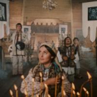 A group of people inside a wooden structure, possibly a church or ceremonial hall, dressed in traditional attire, holding candles amidst smoke, suggesting a religious or cultural ceremony.