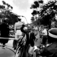 A historical scene featuring a military officer in a white uniform standing in an open-top vehicle, holding a ceremonial staff, surrounded by a crowd, with flags and trees in the background.