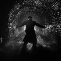 A person stands in a dark, wet tunnel with brick walls, facing away from the camera, with arms outstretched touching the tunnel walls on either side.