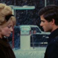 Two individuals standing outside in a snowy environment, near a gas station with falling snow creating a wintry scene.